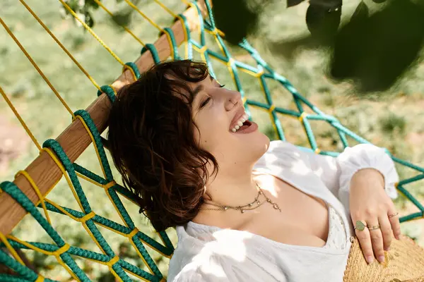 A young woman in a white dress and straw hat laughs joyfully while lounging in a hammock in a summer garden. — Stock Photo