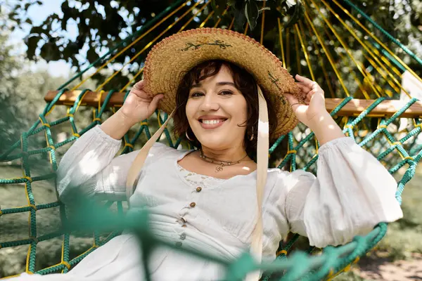 A brunette woman relaxes in a hammock, wearing a white dress and a straw hat, enjoying the warmth of a summer day. — Stock Photo