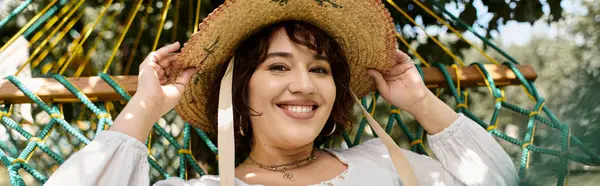 Uma mulher morena em um vestido branco e chapéu de palha sorri enquanto relaxa em uma rede em um dia de verão. — Fotografia de Stock