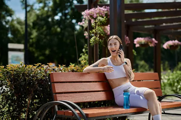 Eine junge Frau in Aktivkleidung sitzt auf einer Parkbank und lacht nach dem Training am Telefon. — Stock Photo