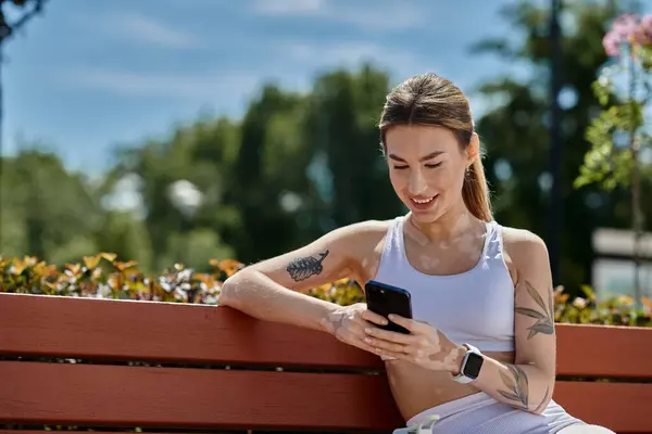 A young woman with vitiligo, wearing activewear, sits on a bench in a park, smiling as she checks her phone. — Stock Photo