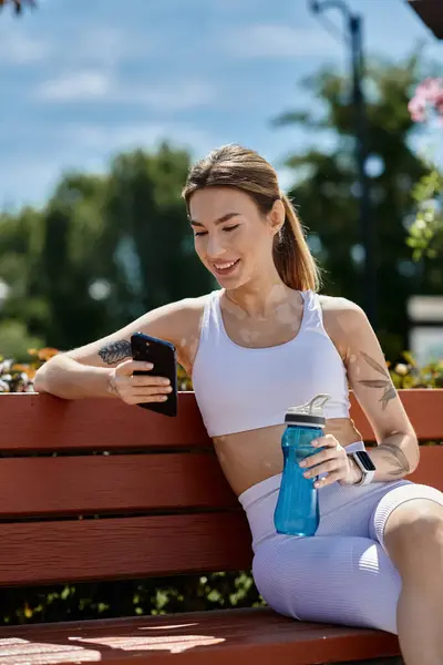 Una joven con vitiligo, en ropa deportiva se sienta en un banco del parque, revisando su teléfono y sosteniendo una botella de agua. — Stock Photo