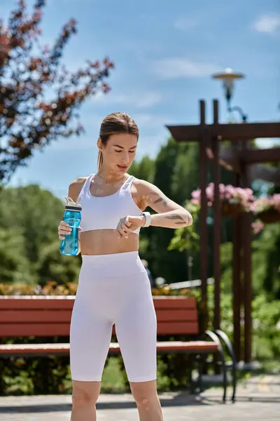 Une jeune femme avec vitiligo vérifie sa montre après une séance d'entraînement dans un parc, tenant une bouteille d'eau. — Photo de stock