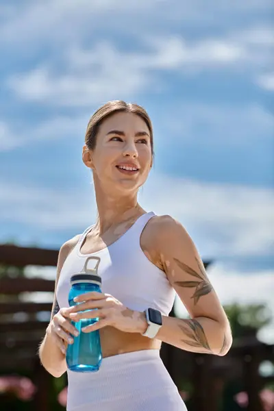 A young woman in activewear, with vitiligo, smiles and holds a water bottle while working out in a park. — Stock Photo
