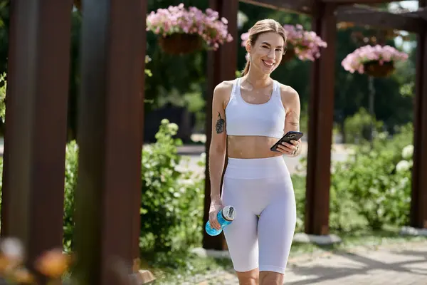 A young woman in white activewear walks through a park, smiling and holding a water bottle and her phone. — Stock Photo