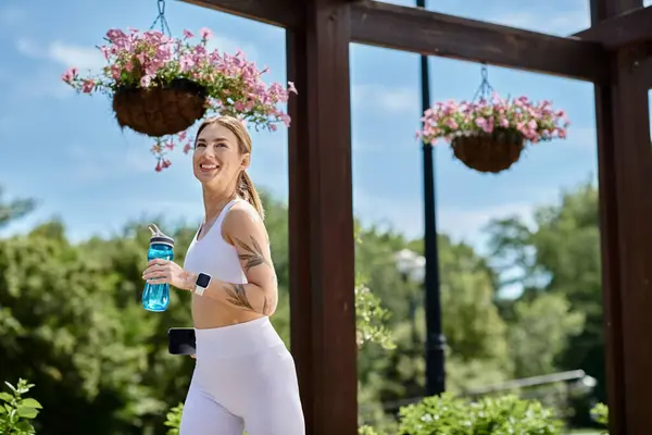 A young woman in white activewear jogs through a park, smiling and holding a water bottle. — Stock Photo