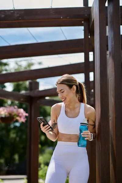 A young woman in white activewear smiles as she checks her phone after a workout, holding a water bottle. — Stock Photo