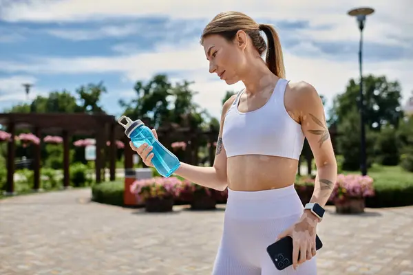 Uma jovem em activewear faz uma pausa de seu treino em um parque, segurando uma garrafa de água na mão. — Stock Photo