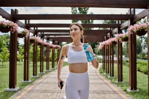 A young woman with vitiligo runs through a park pergola, holding a water bottle and a phone. — Stock Photo