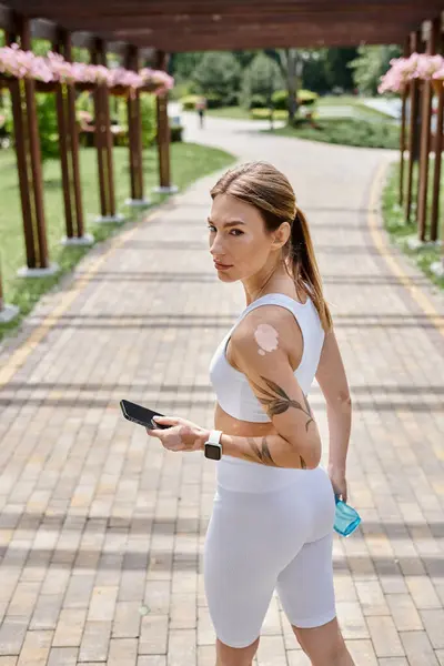 A young woman with vitiligo wears white activewear and a smartwatch as she walks through a park, holding a phone and a water bottle. — Stock Photo