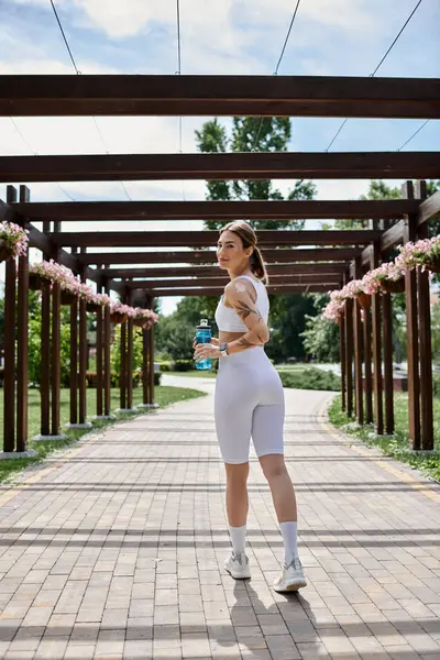 A young sportswoman, dressed in white activewear, walks along a brick path under a wooden pergola, holding a water bottle. — Stock Photo