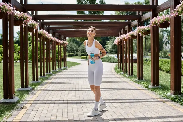 A young woman in white athletic wear walks through a park, smiling with a water bottle in her hand. — Stock Photo