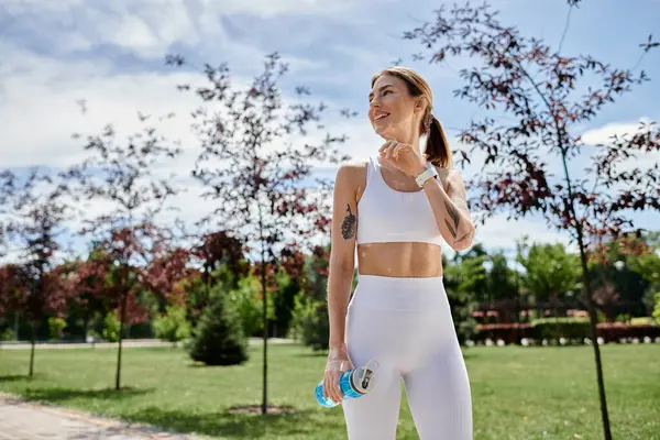 A young woman in white activewear smiles while holding a water bottle after exercising outdoors in a park. — Stock Photo