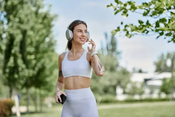 Una giovane donna con la vitiligine, indossando abbigliamento attivo e cuffie, cammina attraverso un parco soleggiato, godendo il suo allenamento. — Foto stock