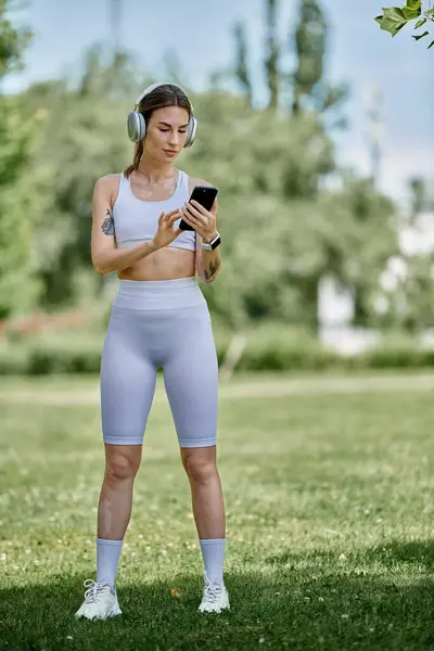 A young woman in activewear, with vitiligo, stands in a park, looking at her phone while wearing headphones. — Stock Photo