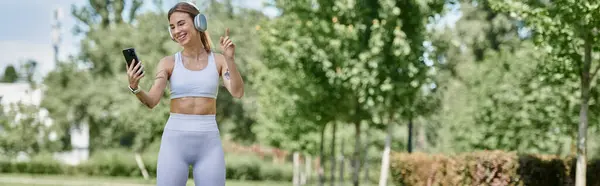 A young woman with vitiligo dances joyfully in a park, wearing activewear and headphones, while holding her phone. — Stock Photo