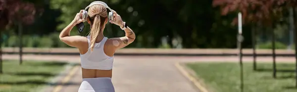 A young woman in athletic wear adjusts her headphones as she walks through a park. — Stock Photo