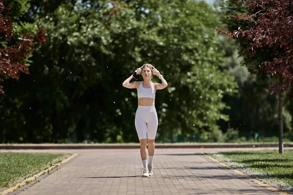 A young woman with vitiligo in white activewear walks on a paved path in a park with trees and green grass on either side. — Stock Photo
