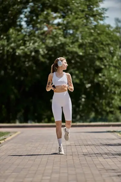 Una giovane donna in activewear bianco corre lungo un sentiero di mattoni in un parco, cuffie accese, telefono in mano, guardando avanti. — Foto stock