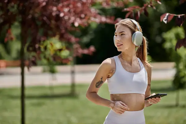 A young woman with vitiligo works out outdoors in a park while listening to music and holding her phone. — Stock Photo
