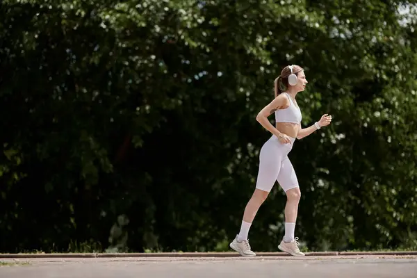A young woman with vitiligo runs through a park, wearing headphones and a white athletic outfit. — Stock Photo
