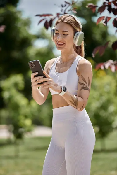 Una joven en ropa deportiva sonríe brillantemente mientras mira su teléfono, los auriculares puestos, durante un descanso de su entrenamiento al aire libre. — Stock Photo