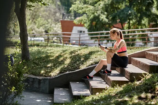 A young woman with vitiligo, wearing a pink sports bra and black shorts, sits on park steps, scrolling on her phone, after a workout. — Stock Photo