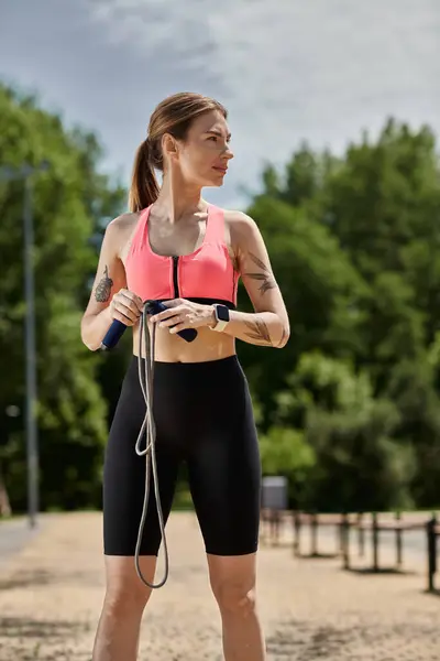 A young woman with vitiligo, wearing a pink sports bra and black shorts, stands outdoors in a park holding a jump rope. — Stock Photo