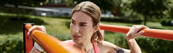 Una joven con vitiligo, vistiendo una blusa rosa y ropa deportiva, realiza un ejercicio pull-up al aire libre en un parque. - foto de stock