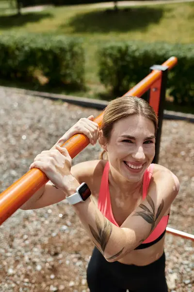 A young woman with vitiligo, wearing a pink crop top, smiles as she works out on a horizontal bar in a park. — Stock Photo