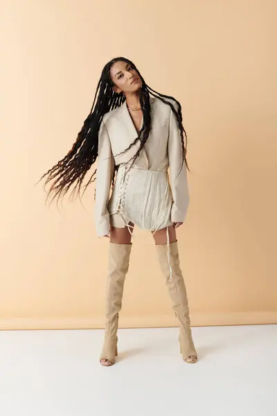 A young African American woman with long braids poses in a stylish outfit, wearing a white blazer, white skirt and knee-high boots. — Stock Photo