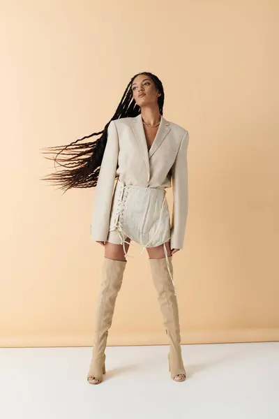 Une jeune femme aux longs cheveux noirs porte un blazer blanc, une jupe avec des détails en dentelle, et des bottes au-dessus du genou dans un studio. — Photo de stock