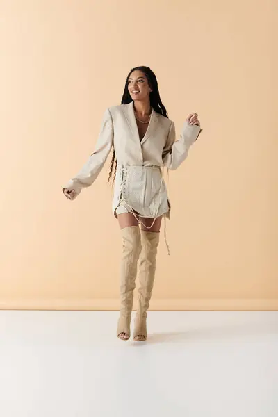 A young woman with long black hair poses in a white blazer, skirt, and thigh-high boots against a pale beige background. — Stock Photo