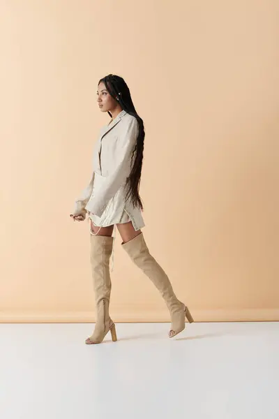 A stylish young woman with long braids walks in a studio, wearing a white jacket and matching skirt and knee-high boots. — Stock Photo