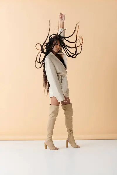 A young woman in stylish attire poses on a beige backdrop. Her long, dark braids flow freely around her. — Stock Photo