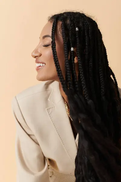 Una joven afroamericana con el pelo largo trenzado sonríe mientras usa una chaqueta de crema sobre un fondo beige. - foto de stock