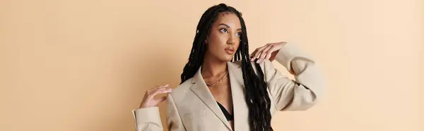 A young woman with long black braids poses confidently in a beige blazer against a neutral backdrop. — Stock Photo