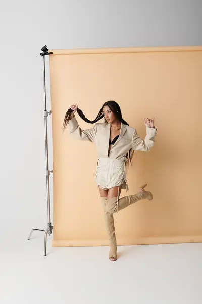 A young African American woman poses against a beige backdrop, wearing a white blazer, a short skirt, and thigh-high boots. — Stock Photo