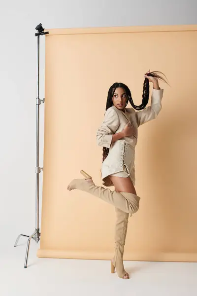 A young Black woman poses in stylish beige attire, playfully holding one of her long braids. The background is a soft, neutral beige. — Stock Photo