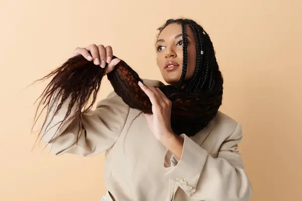 Une jeune femme avec de longues tresses noires pose sur un fond neutre, portant un blazer de couleur claire et une expression réfléchie. — Photo de stock