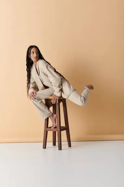 Una mujer con estilo en un traje beige se sienta en un taburete, mirando por encima de su hombro con una expresión segura. - foto de stock