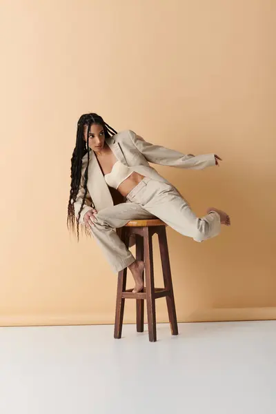 A stylish young woman in a blazer and pants sits on a wooden stool against a beige background. — Stock Photo