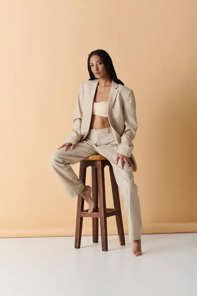 A young woman in beige pantsuit sits on a wooden stool against a pale beige backdrop. — Stock Photo