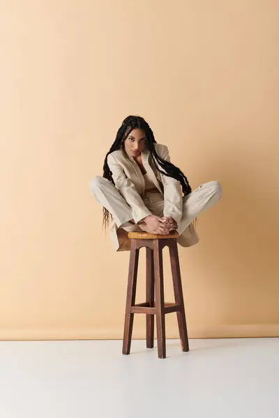 A young woman in a light-colored suit sits cross-legged on a wooden stool against a beige backdrop. — Stock Photo