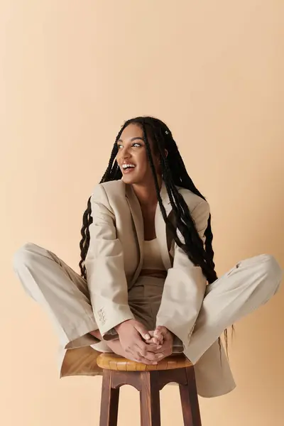 Una joven con trenzas largas se sienta en un taburete de madera con un traje beige sobre un fondo beige silenciado. - foto de stock