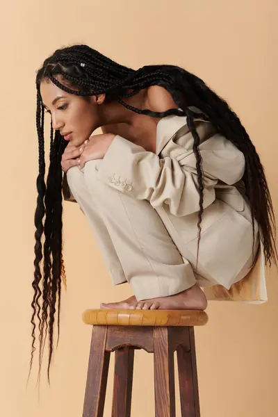 A young woman with long black braids sits on a stool against a beige backdrop, her head bowed in thought. — Stock Photo