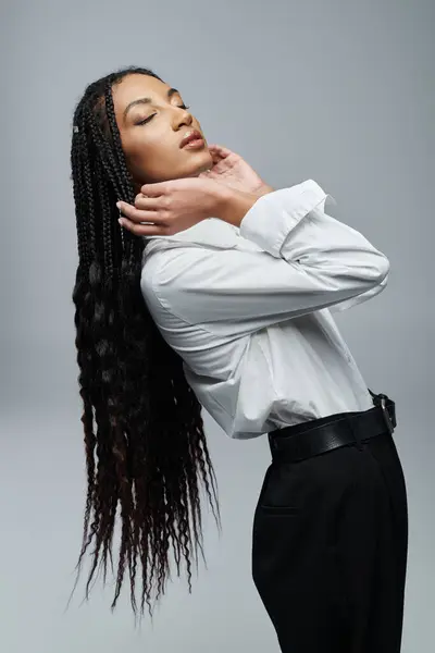 Uma jovem negra com longos cabelos trançados veste uma camisa branca e posa com a cabeça virada para trás contra um pano de fundo cinza.. — Fotografia de Stock