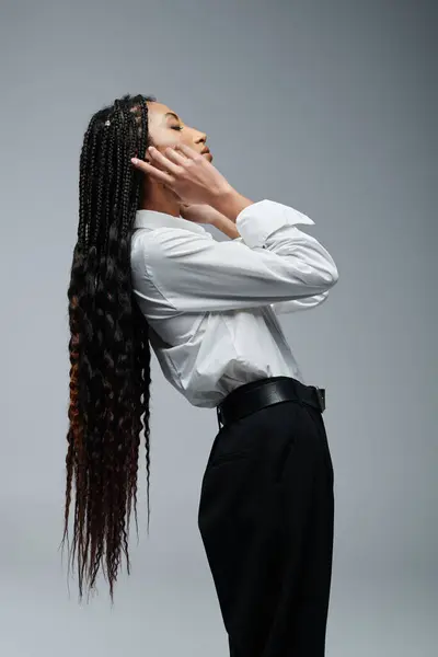 A young woman with long braids poses in a white shirt and black pants against a grey background. — Stock Photo