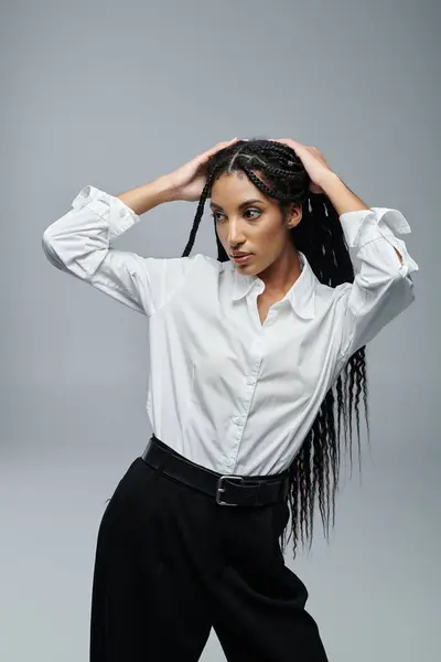 A young woman in a white shirt and black pants poses against a grey backdrop, her hand gently stroking her long braids. — Stock Photo