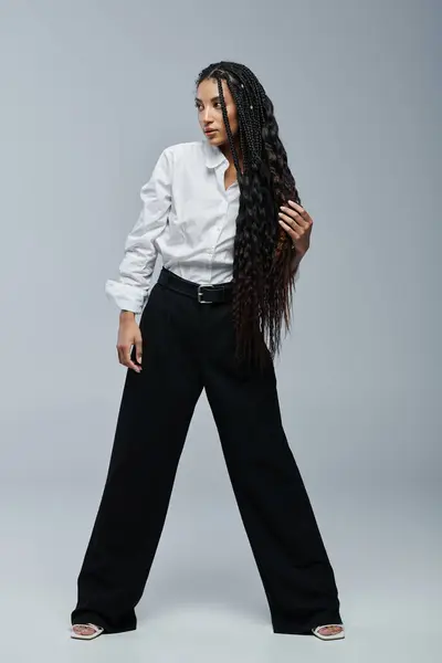 A young black woman with long braided hair poses in a white shirt and black pants against a grey background. — Stock Photo
